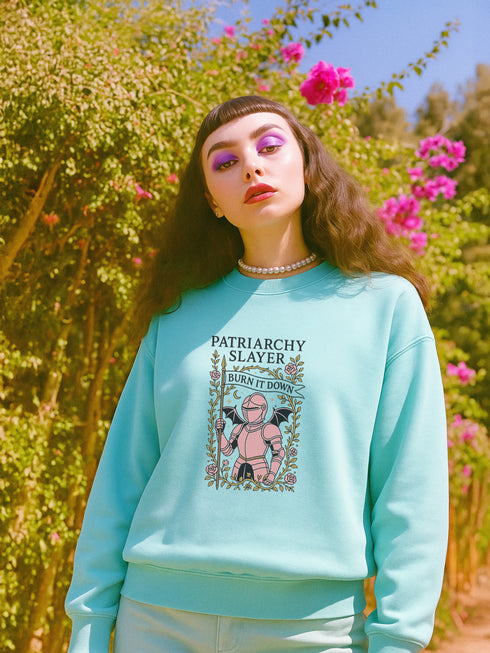 Comfort Colors garment-dyed sweatshirt featuring 'Patriarchy Slayer' design with pink armored knight holding sword and banner reading 'Burn It Down', surrounded by decorative floral border with roses and leaves. Made from 80% ring-spun cotton and 20% polyester with relaxed fit and rolled-forward shoulders. Available in lavender purple and seafoam green colorways, sizes S-3XL unisex, featuring sustainable made-to-order production with OEKO-TEX certified low-impact dyes and zero waste manufacturing process.
